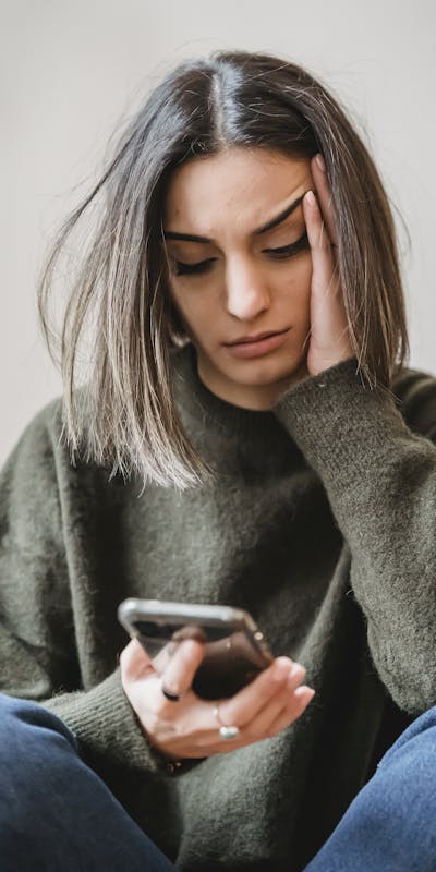 Unhappy female in casual wear leaning on hand and surfing internet on cellphone while sitting in light room near wall at home